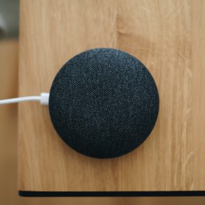 A small, round smart speaker rests on a wooden surface, plugged in with a white cable, near the edge of a tabletop.