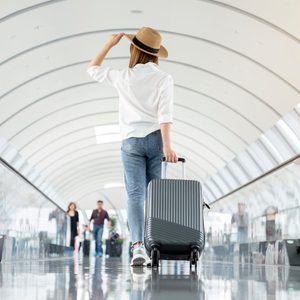 Traveler pulls suitcase while walking through a bright, modern airport terminal, wearing a hat and jeans. Others walk in the background.