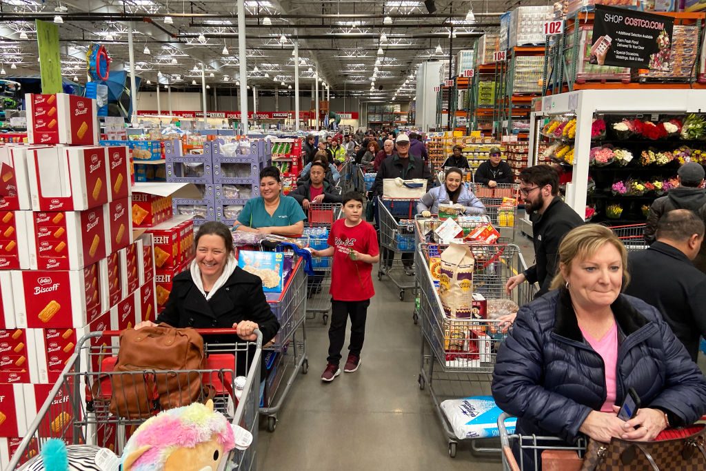Shoppers push carts along crowded store aisle, surrounded by stacked products and floral displays, creating a bustling retail environment.