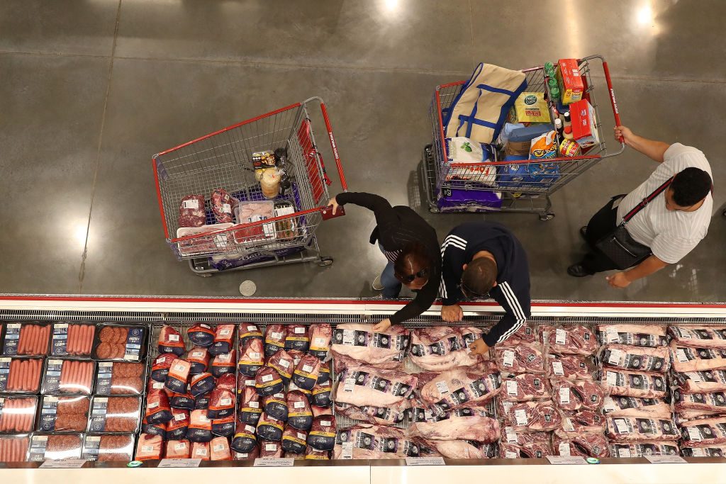 Shoppers examine packaged meat in a grocery store, pushing carts filled with various items on a polished concrete floor.