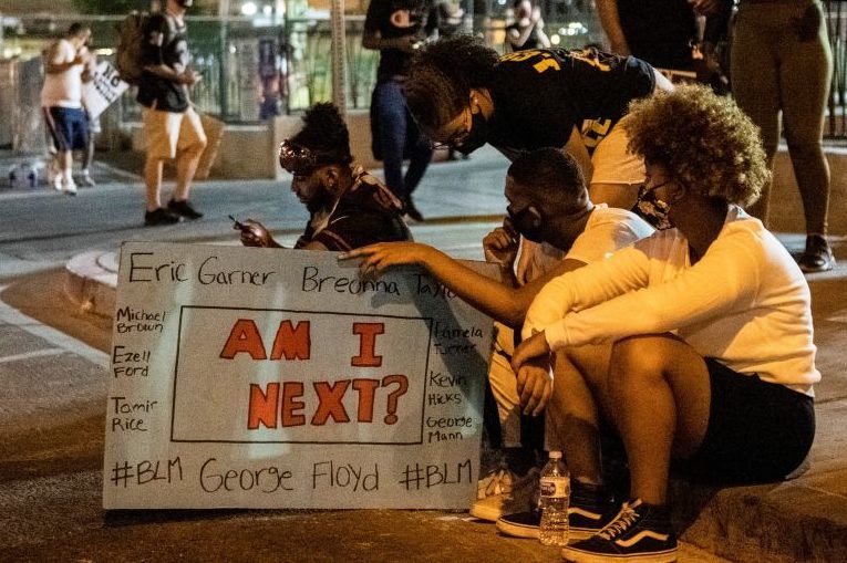 People kneel holding a sign reading "AM I NEXT?" with names and hashtags, sitting on a street at night, other people in the background.