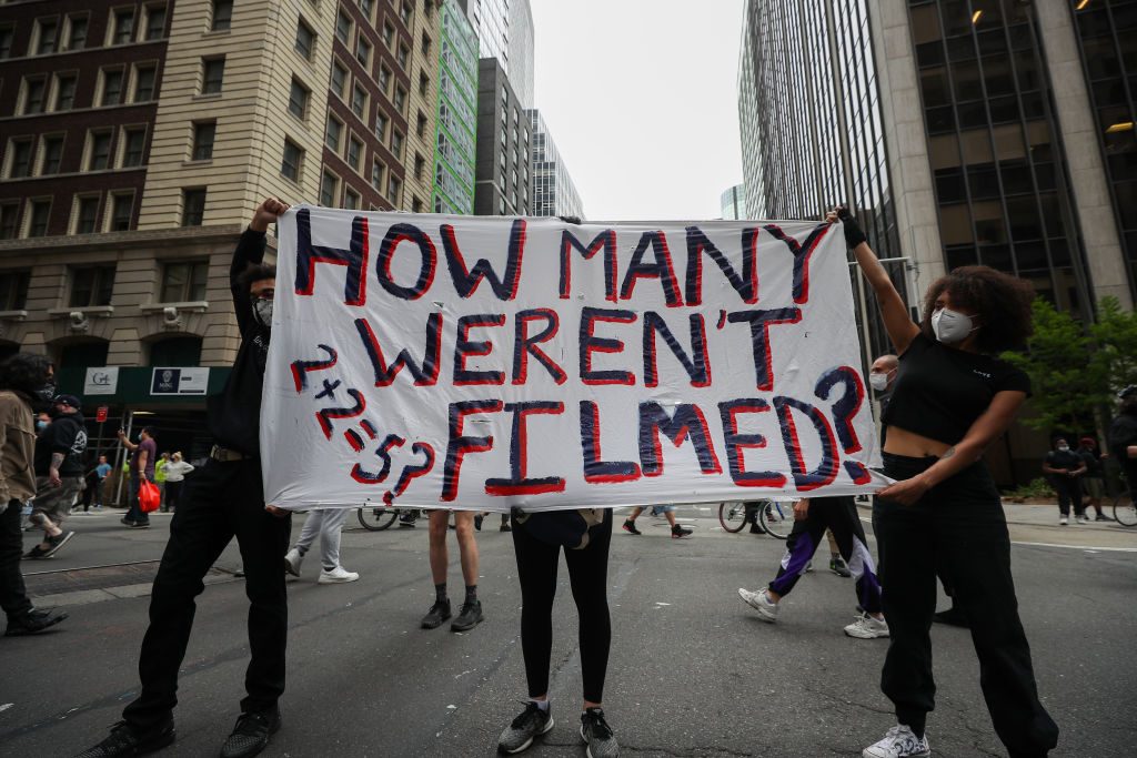 Protesters hold a sign reading "HOW MANY WEREN'T FILMED?" on a city street surrounded by tall buildings and pedestrians.
