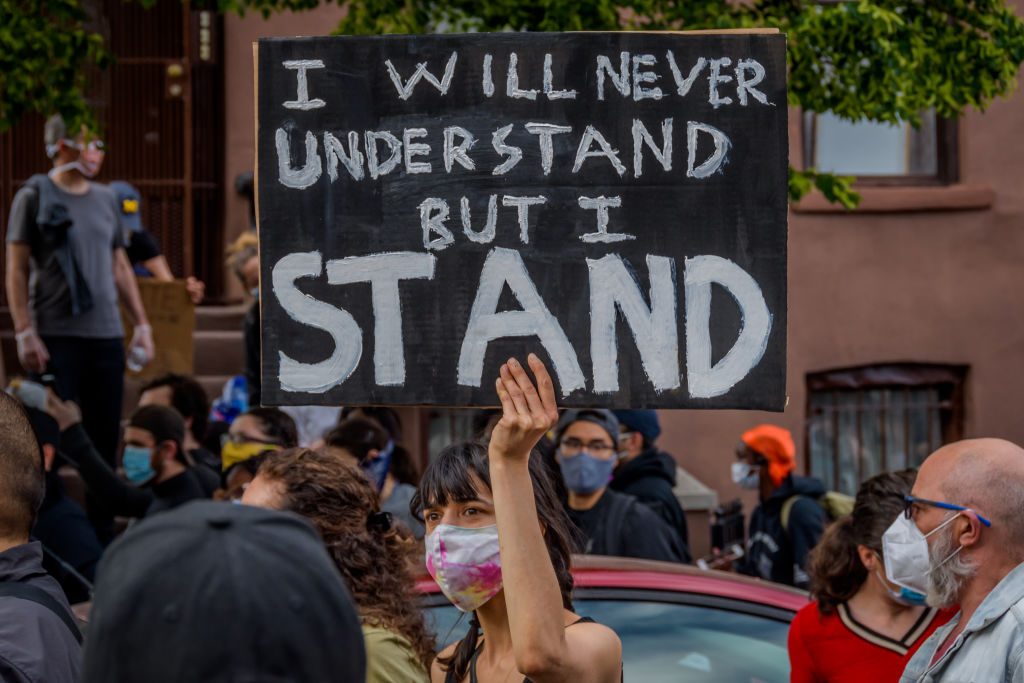 Sign displays "I WILL NEVER UNDERSTAND BUT I STAND" held by person in protest crowd, with people wearing masks around.