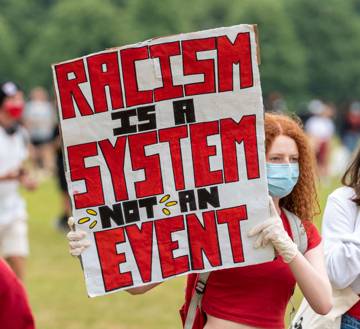 A person holds a sign reading "RACISM IS A SYSTEM NOT AN EVENT" at an outdoor protest with people in the background.