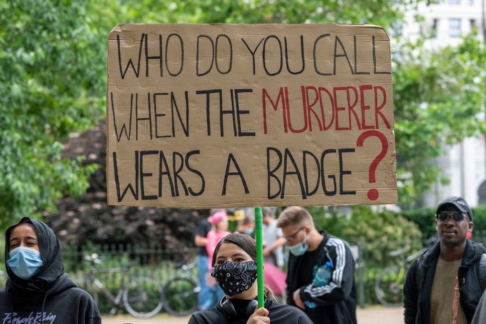 Person holding a sign reading "WHO DO YOU CALL WHEN THE MURDERER WEARS A BADGE?" amidst a crowd in a park setting.