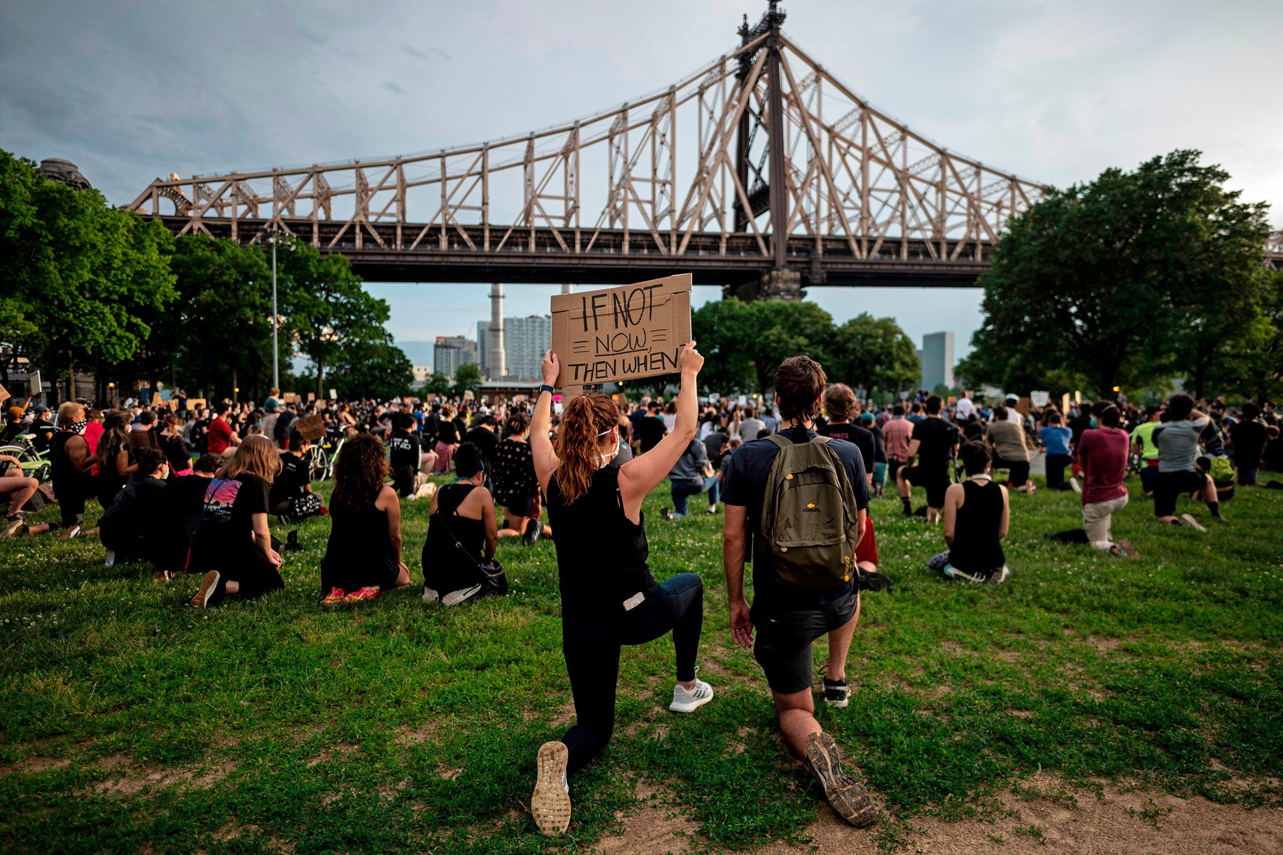Person kneels, holding sign saying "IF NOT NOW THEN WHEN," amidst a crowd kneeling on grass, with a bridge and skyline in the background.