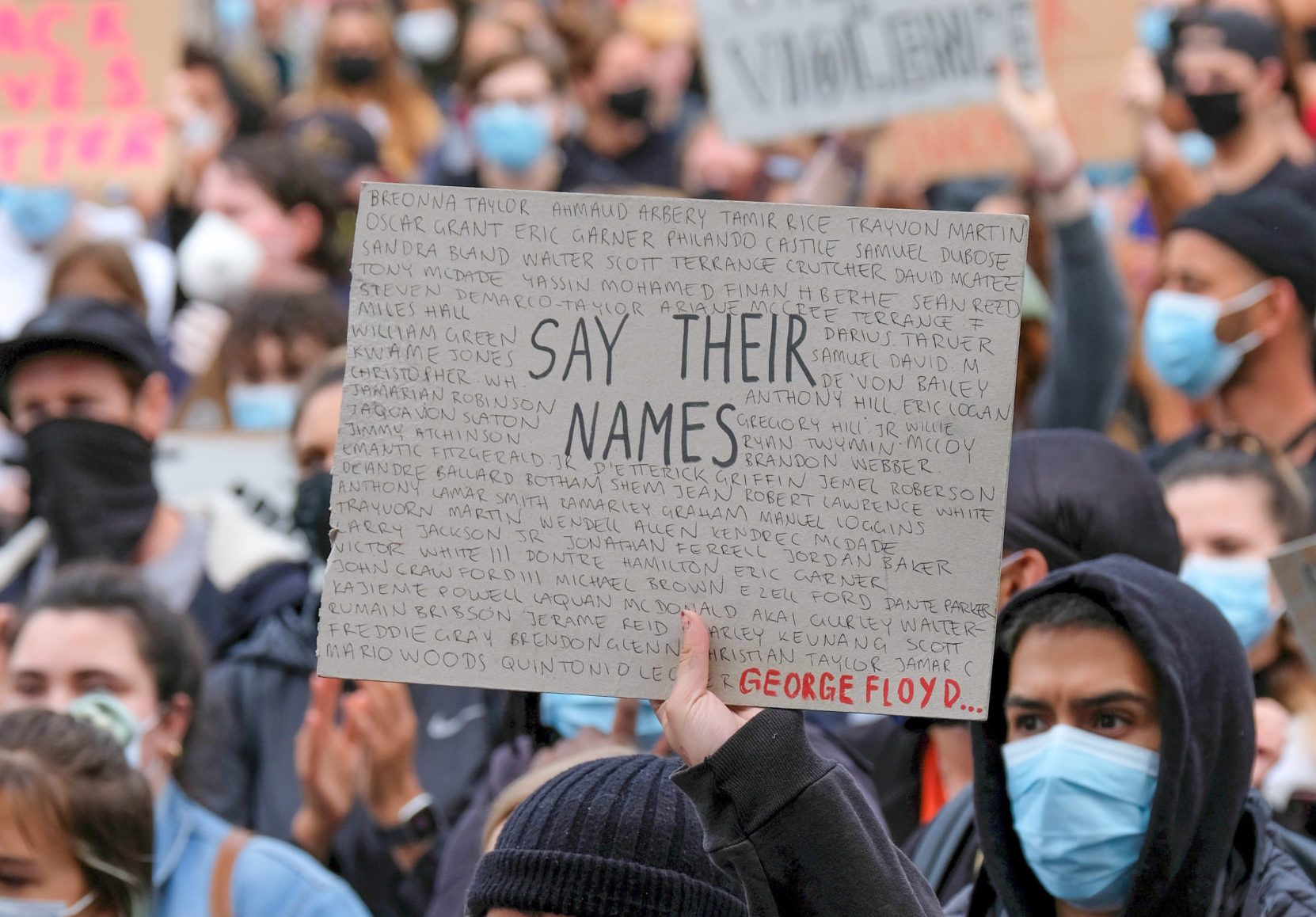 Person holds a sign saying "Say Their Names" with handwritten names, amidst a crowd wearing masks at a public protest.
