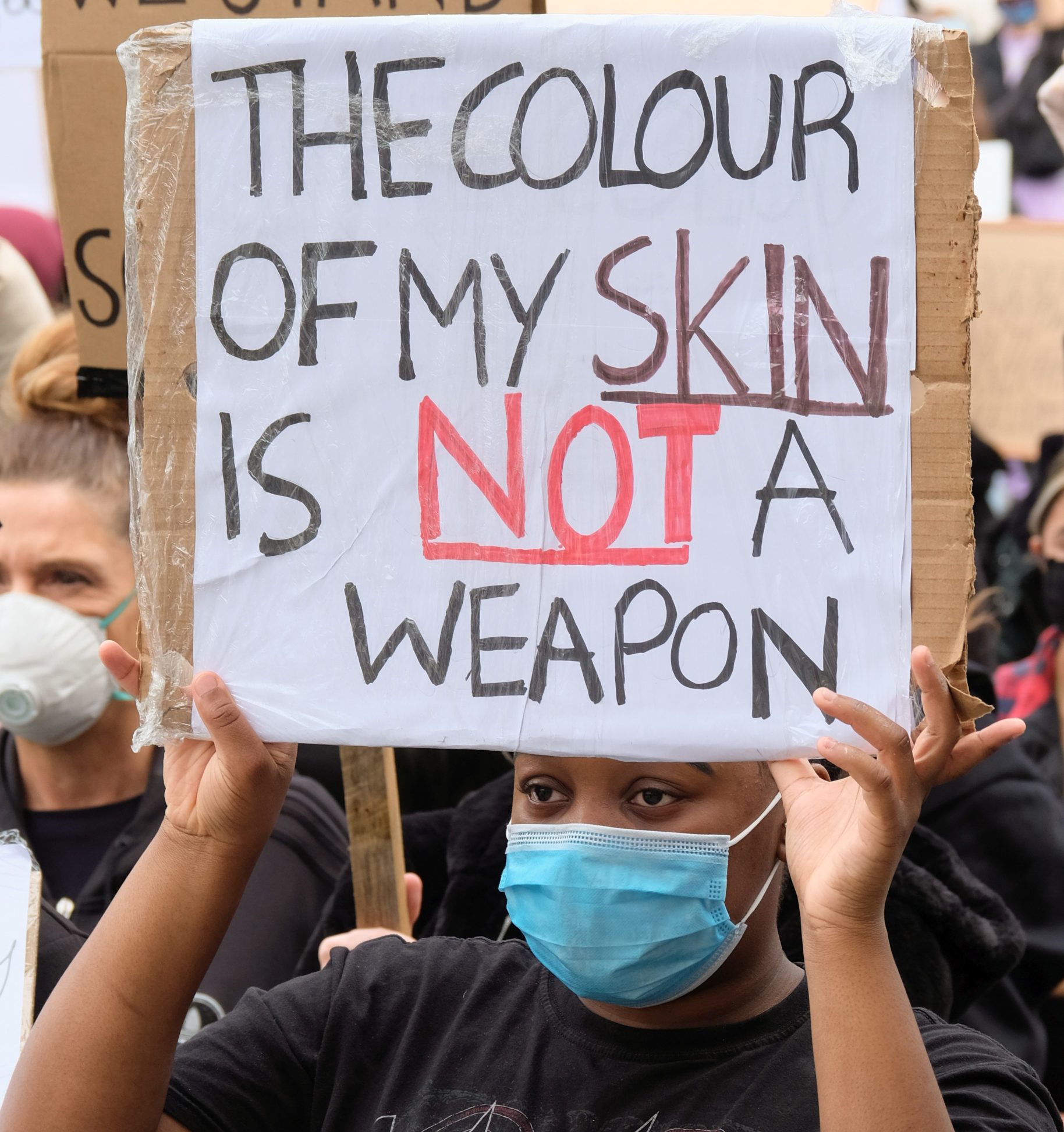 Person holds protest sign saying "THE COLOUR OF MY SKIN IS NOT A WEAPON" in a crowd, wearing a blue mask.