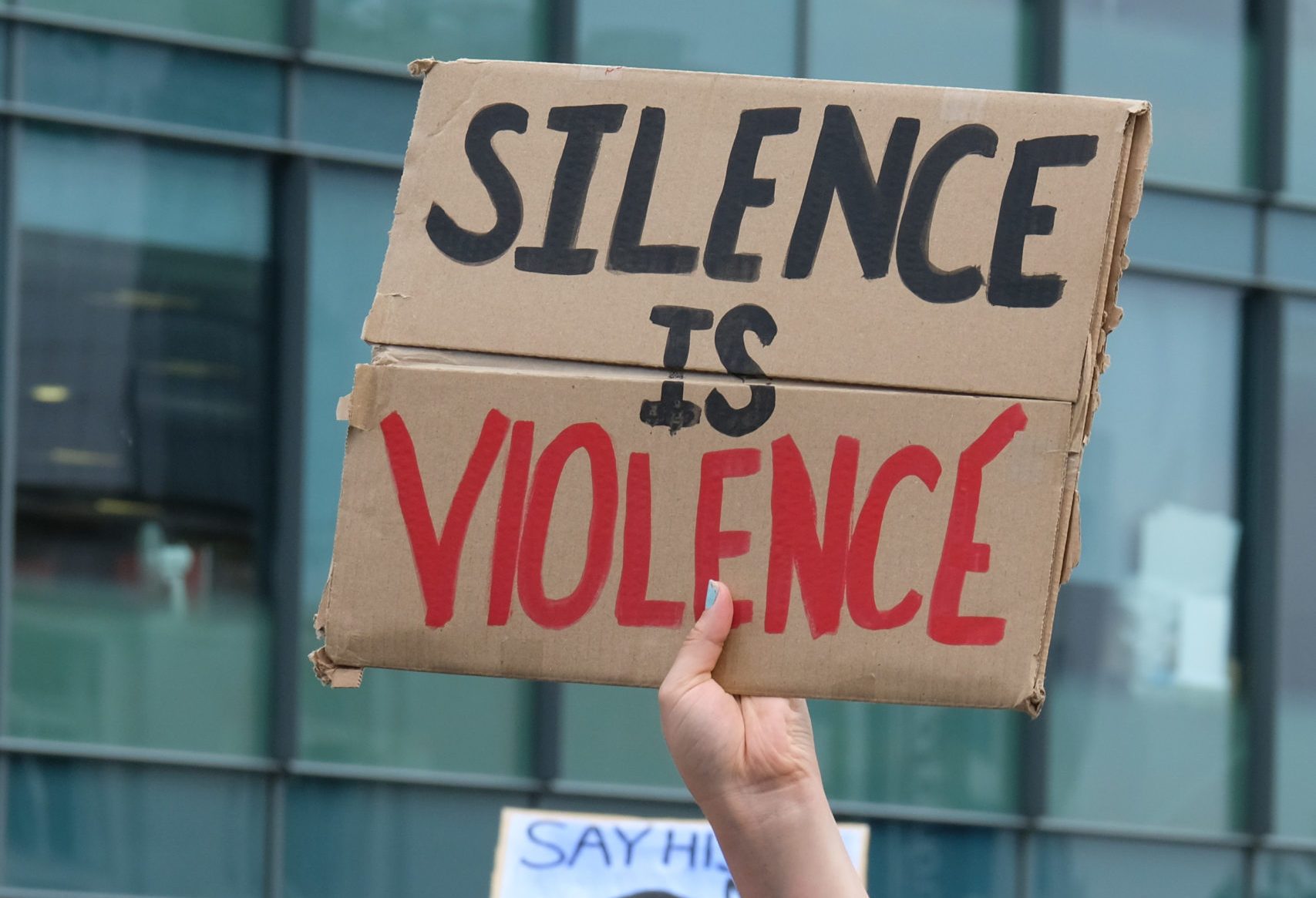 Hand holds cardboard sign reading "SILENCE IS VIOLENCE" against a backdrop of glass-paneled buildings.