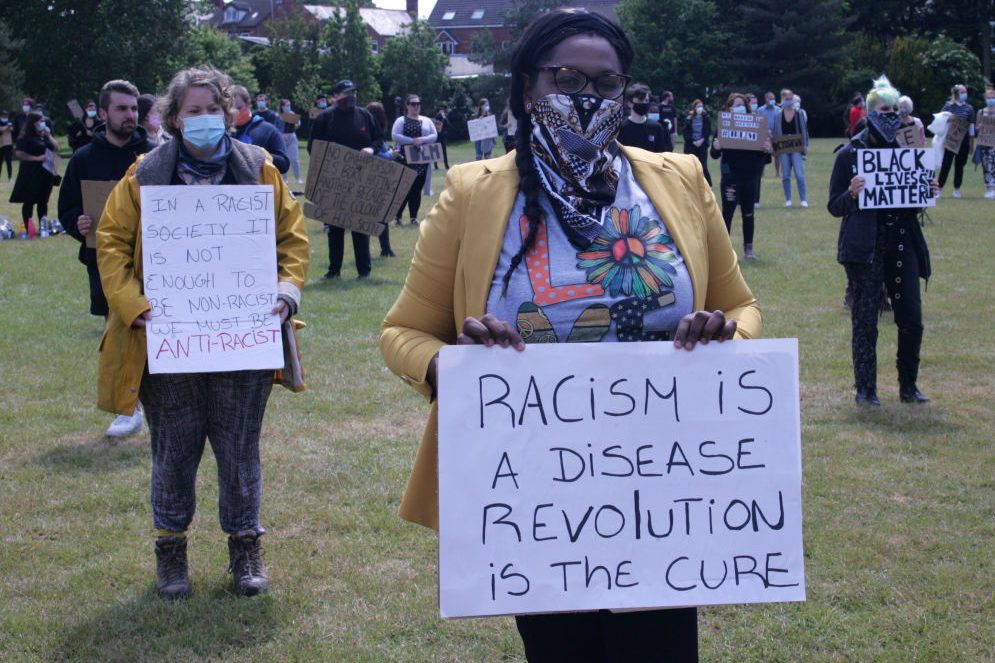 Individuals hold signs at a protest on a grassy area. Sign text includes: "Racism is a disease, revolution is the cure" and "we must be anti-racist."