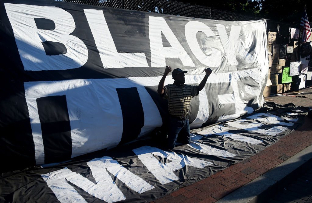 Banner displays "BLACK LIVES MATTER"; man kneels with raised fists in front of it, surrounded by protest signs on a brick pathway.