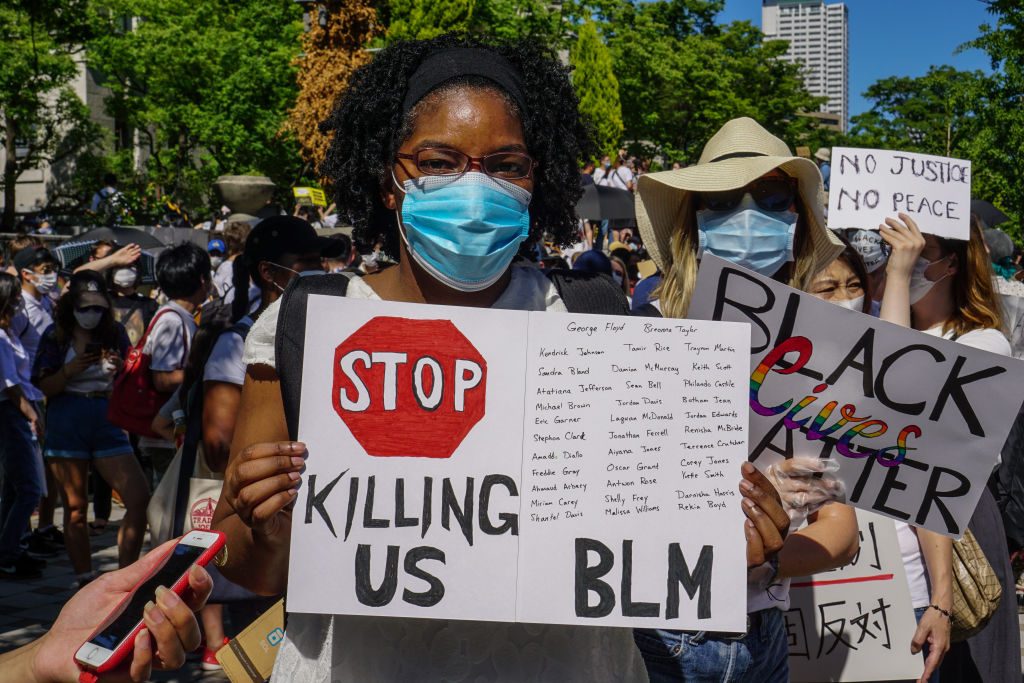 Protester holds a sign reading "STOP KILLING US BLM" with names written above, surrounded by a crowd with signs, outdoors.