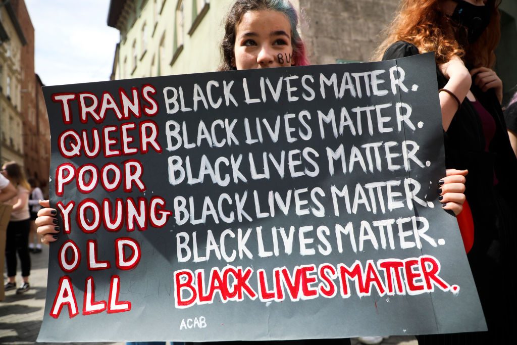 Person holding a sign listing "TRANS, QUEER, POOR, YOUNG, OLD, ALL" with "BLACK LIVES MATTER" alongside each. Background shows people and buildings.