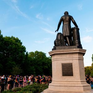 A statue stands prominently on a pedestal, surrounded by a crowd of people in a park setting with lush green trees and a clear sky.