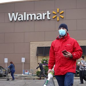 A person wearing a red jacket and mask walks, holding a phone, outside a Walmart store. Shoppers with carts are in the background.