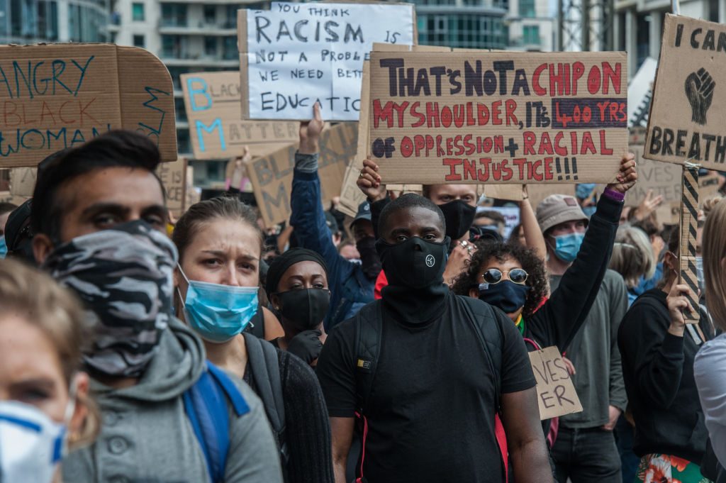 Crowd holds protest signs about racial injustice, wearing masks, in an urban setting. One sign reads: "That's not a chip on my shoulder..."