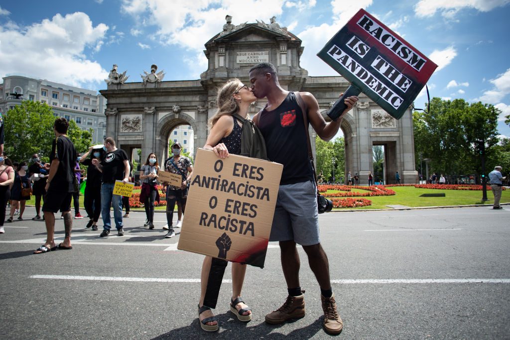 Protesters kiss while holding signs reading "Racism is the pandemic" and "O eres antirracista o eres racista" near a historic arch in a city park.