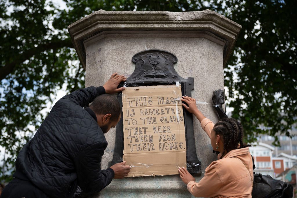 People attach cardboard sign to statue base; sign reads, "THIS PLAQUE IS DEDICATED TO THE SLAVES THAT WERE TAKEN FROM THEIR HOMES." Trees and buildings in background.