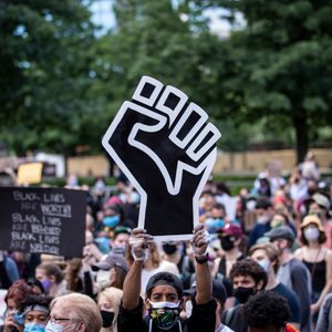 A person holds a large black fist sign above a diverse crowd, surrounded by protestors with signs in a park setting.