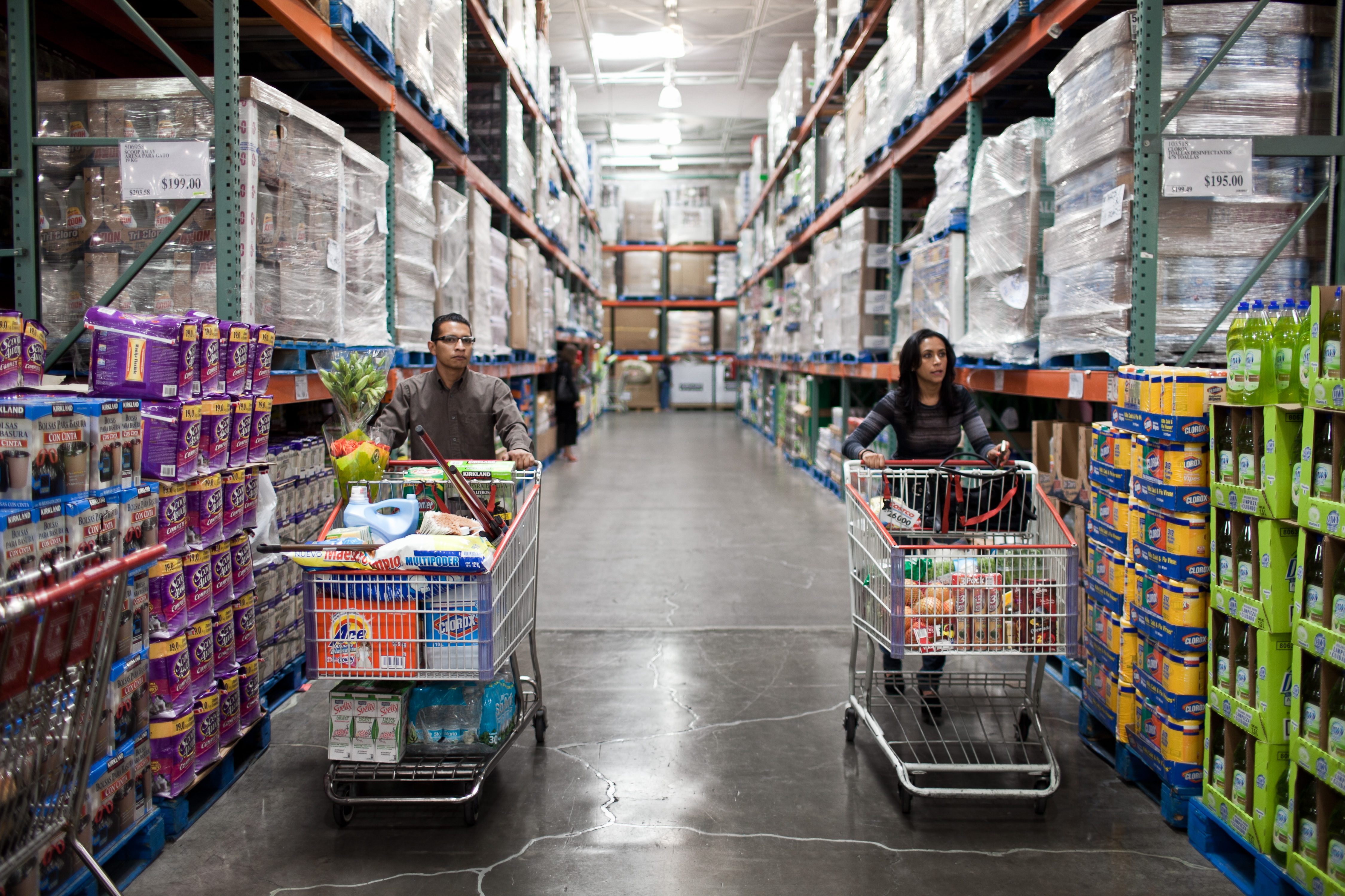 Two shoppers push full carts through a large warehouse store aisle lined with shelves of bulk products.