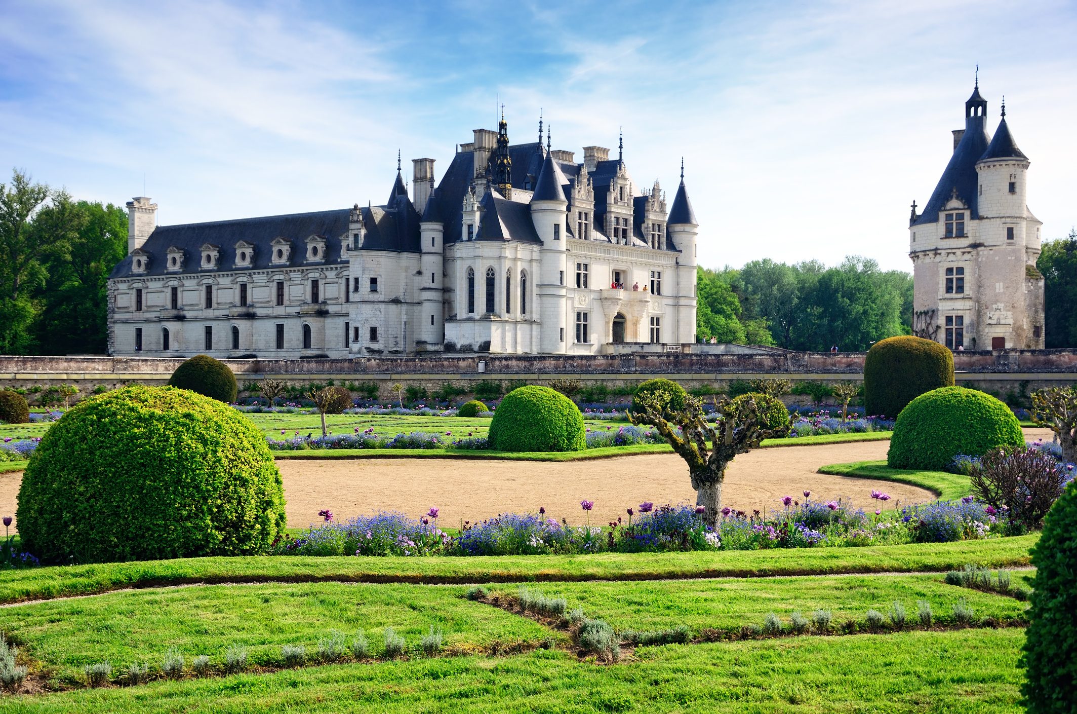 Castle stands elegantly with turrets; viewed from a manicured garden with geometric hedges and vibrant flowers, under a clear blue sky.