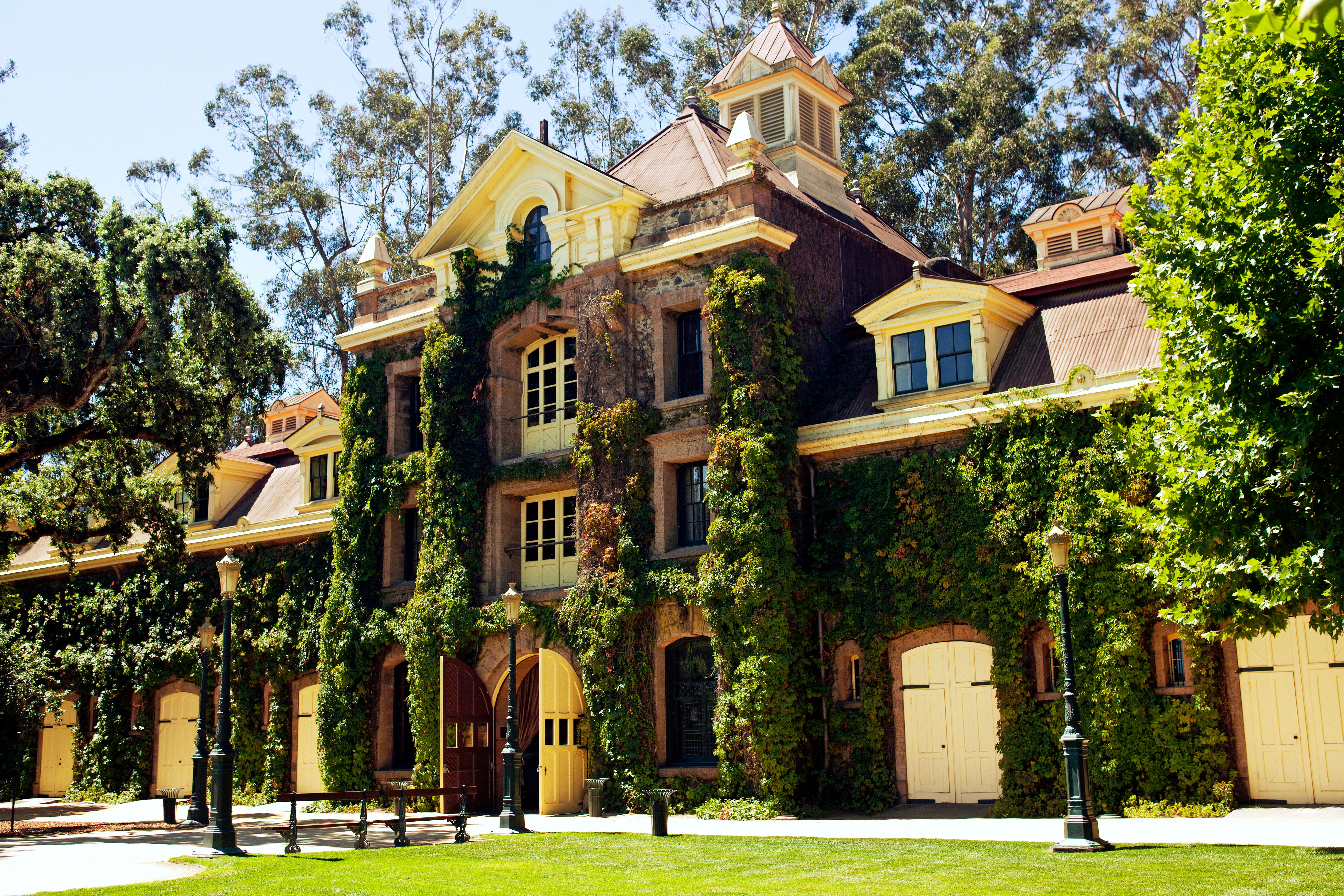 A historic building, covered in ivy, stands amidst trees and bright sunlight, with a lush green lawn in the foreground.