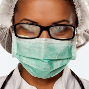Person wearing fogged glasses and a surgical mask, dressed in medical attire, with a hair cover, suggesting a healthcare or lab setting.