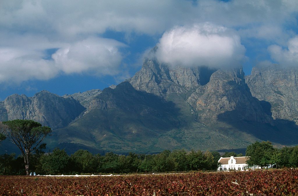 Mountain looms with clouds enveloping peaks; a vineyard stretches below. A white house stands amidst green trees, under a blue sky with scattered clouds.