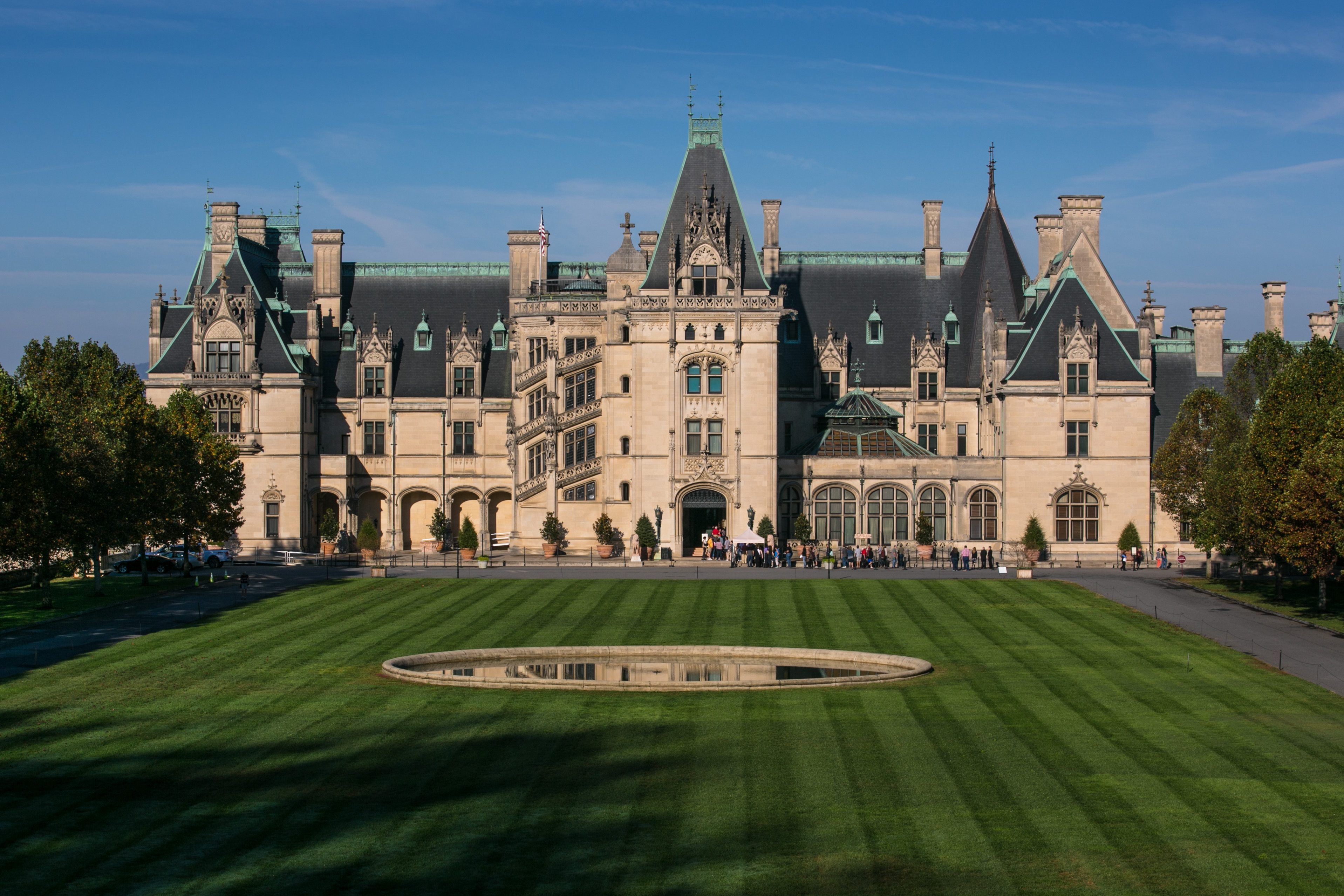 A large, ornate mansion stands in front of a neatly trimmed lawn with trees, under a clear blue sky.