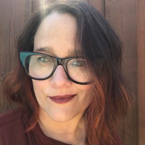 A person is smiling slightly, wearing glasses with sunlight behind them, set against a wooden fence background.