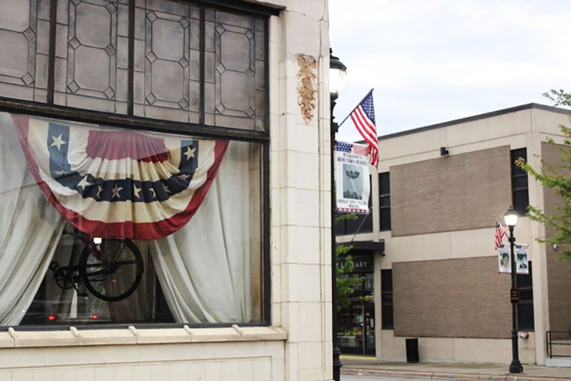 A window displays a patriotic banner inside a building with brick and stucco walls. Nearby, American flags decorate a street scene with lampposts.