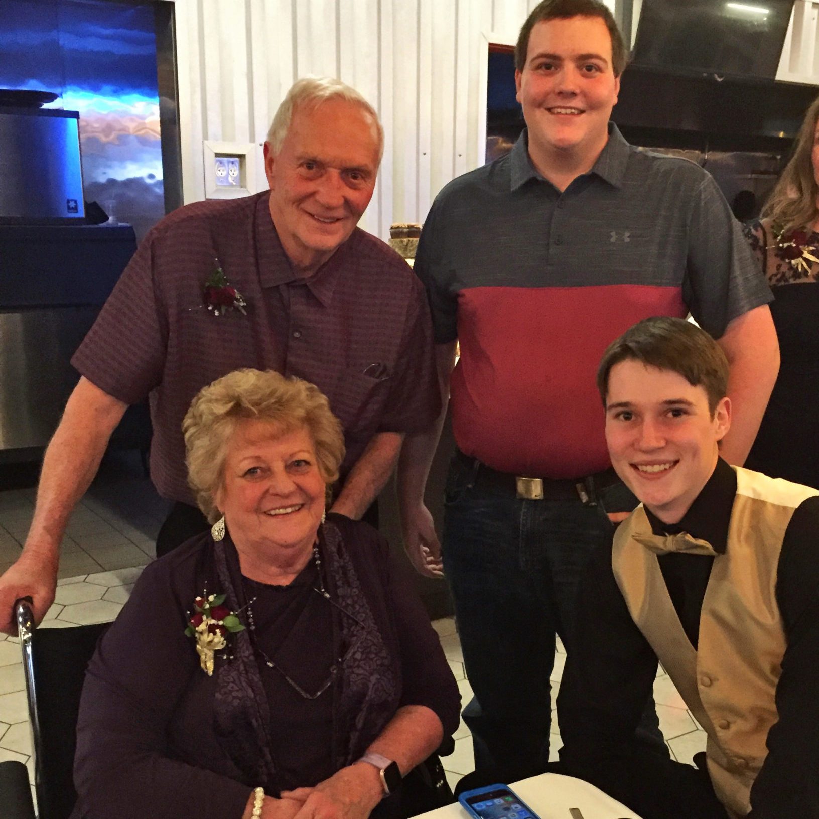 Four people smile in a warmly lit room; two stand behind, two sit in front. A phone lies on the table.