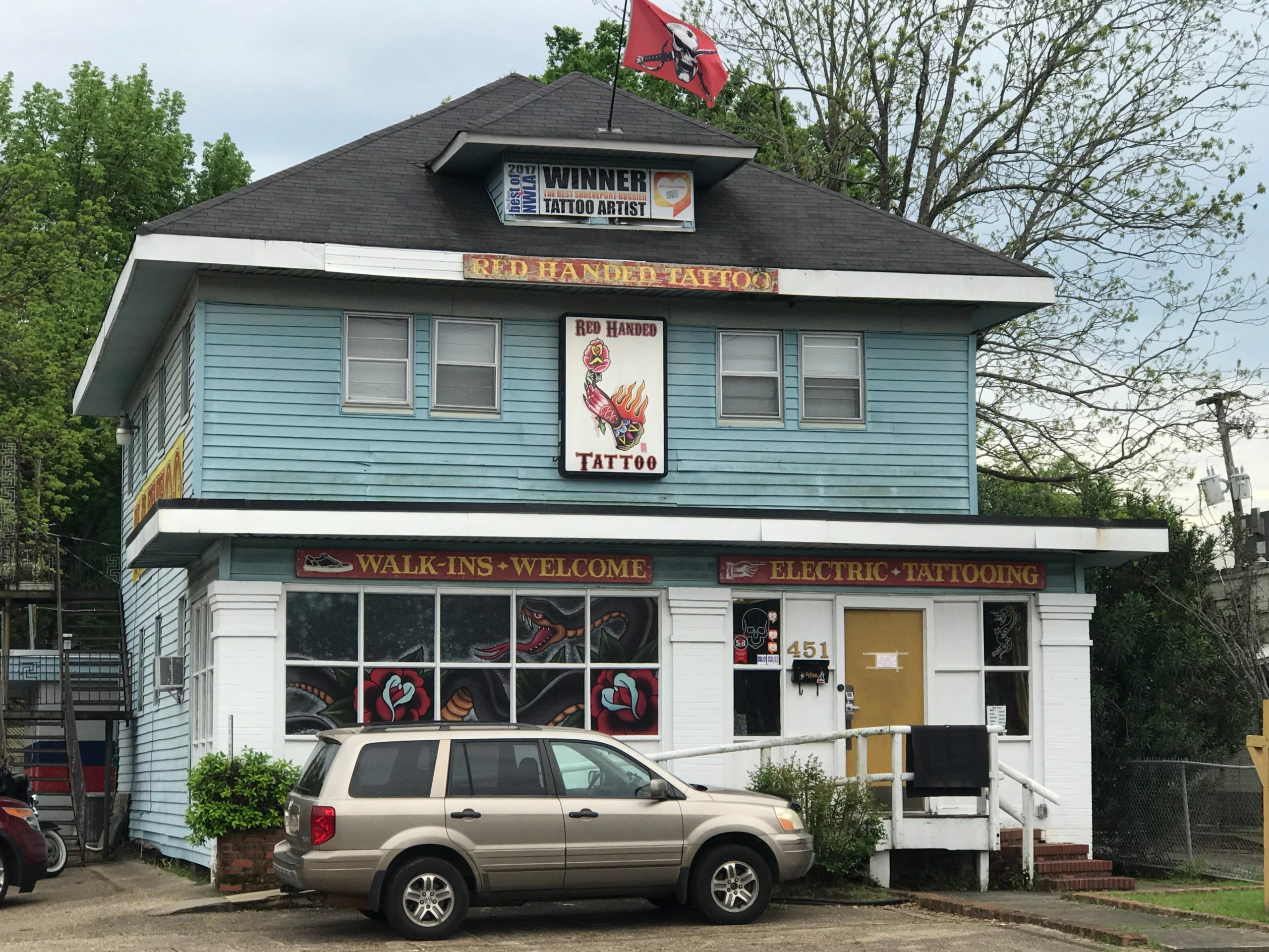 Tattoo shop displays artwork while cars park outside; signs read "WINNER TATTOO ARTIST," "RED HANDED TATTOO," and "WALK-INS WELCOME ELECTRIC TATTOOING."