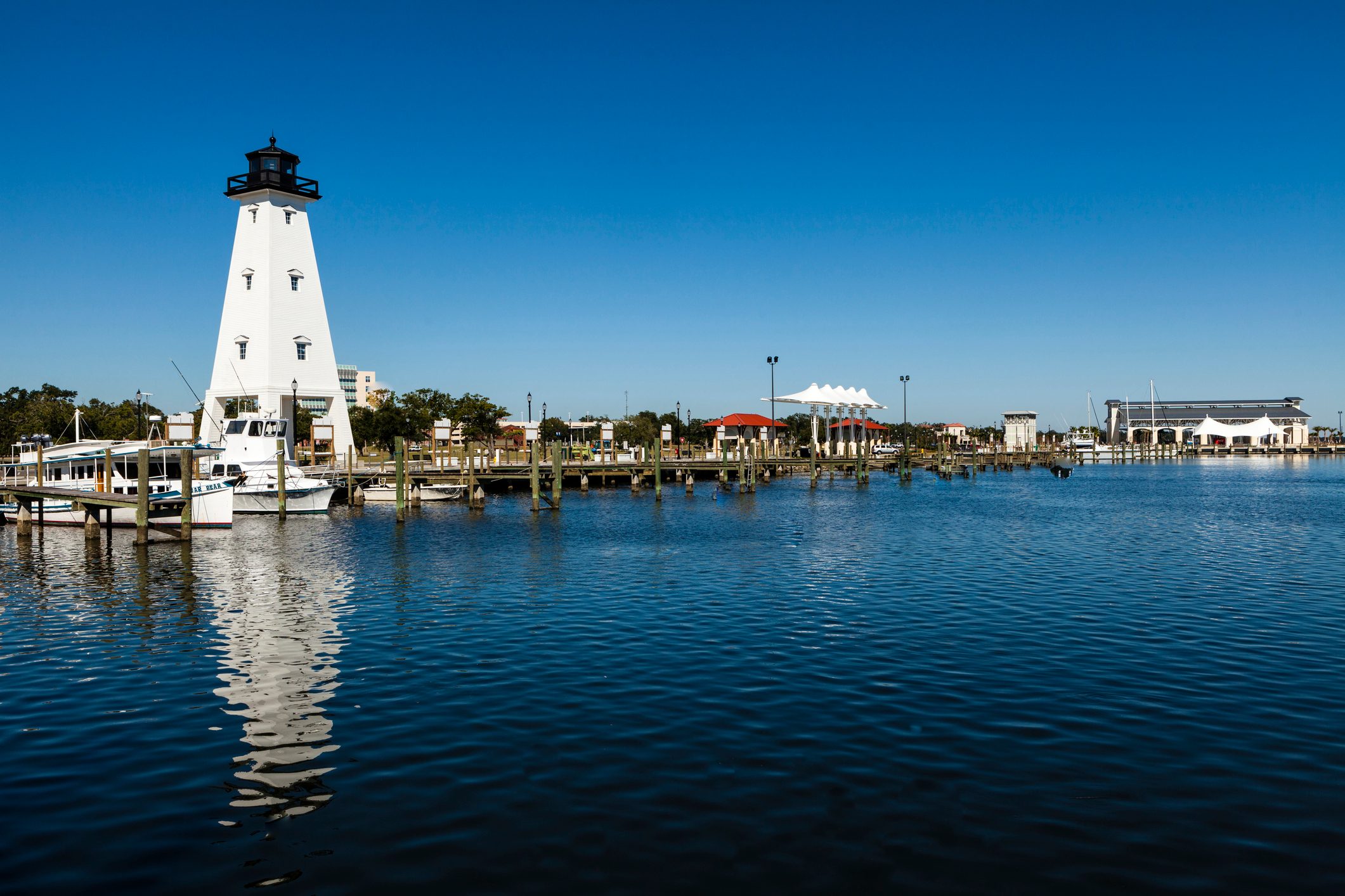 A white lighthouse stands by the waterfront, reflected in calm blue water, surrounded by docks and boats under a clear sky.