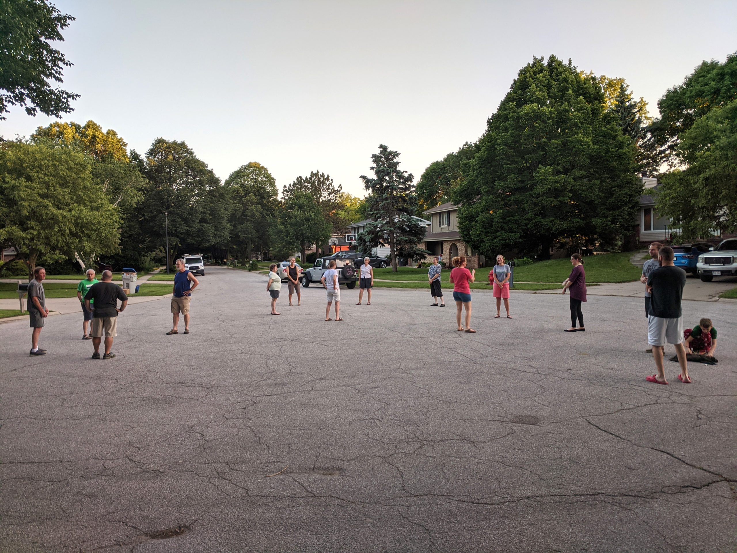 People stand chatting in small groups on a residential street, surrounded by parked cars and lush green trees under a clear sky.
