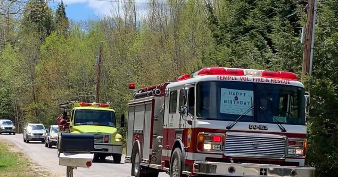 Red fire truck displays "HAPPY BIRTHDAY" sign while driving in a procession on a tree-lined rural road, followed by other vehicles.