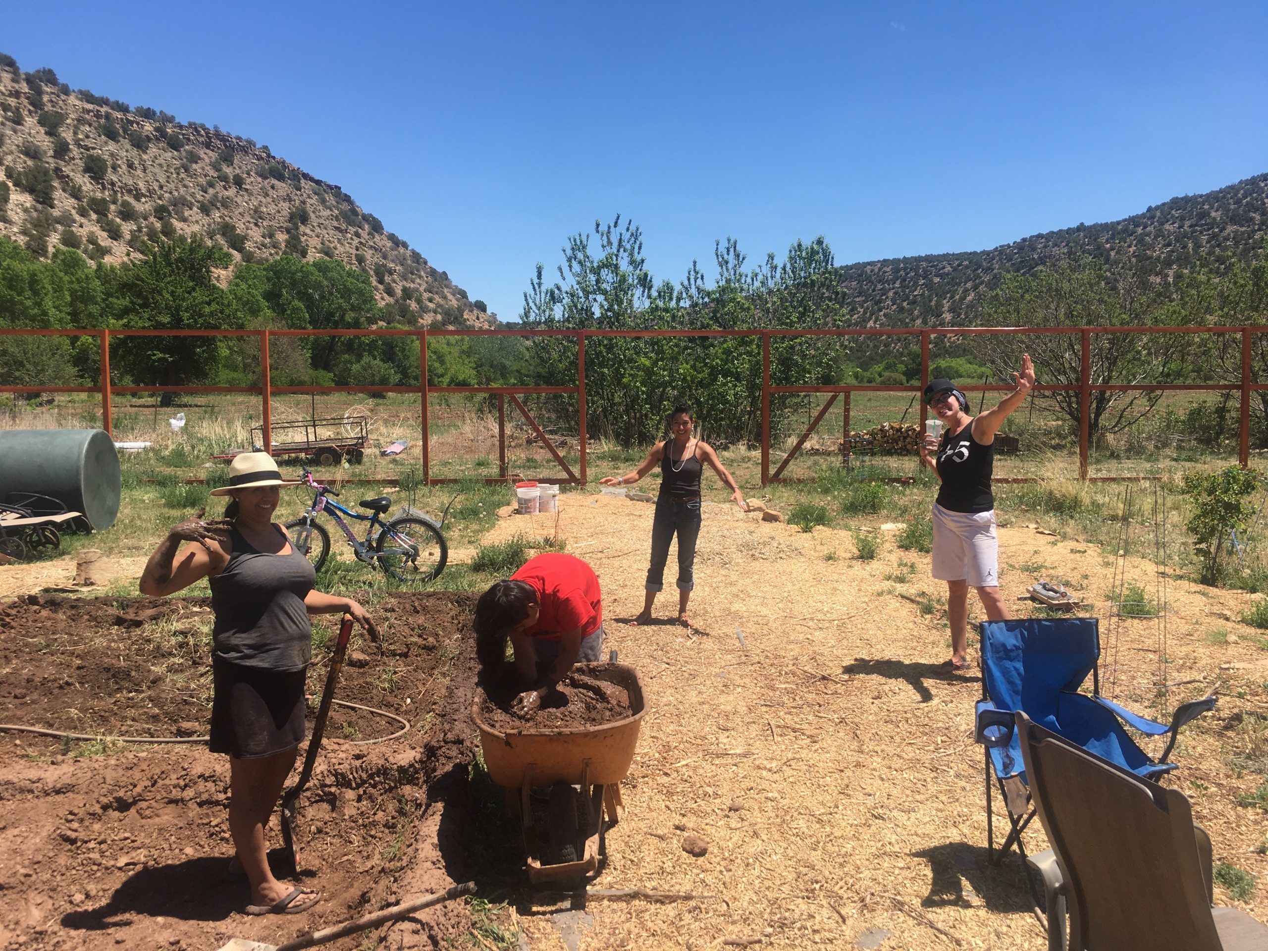 People garden under a clear sky; one shovels soil, another gathers dirt in a wheelbarrow. Fenced area with mountains and greenery surrounds them.