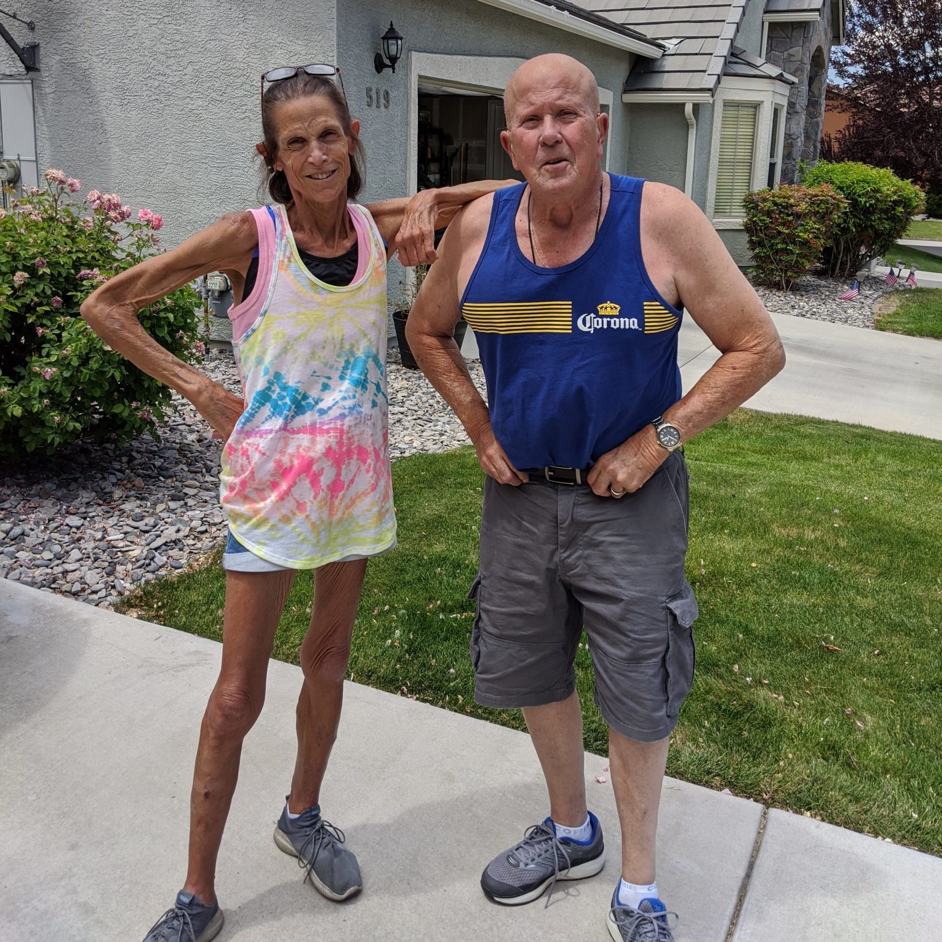 Two people stand confidently on a sidewalk, with a house and garden in the background.