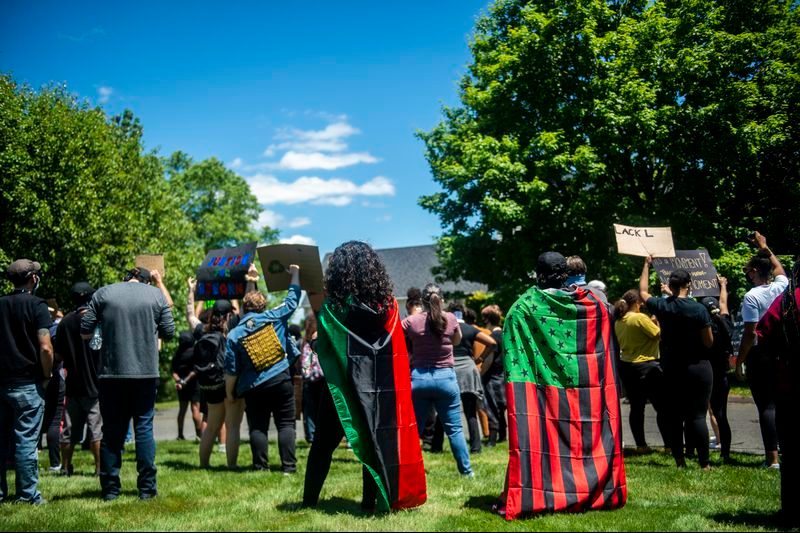 Protesters stand holding signs, wearing colorful flags as capes, in a park setting surrounded by vivid green trees and a clear blue sky.