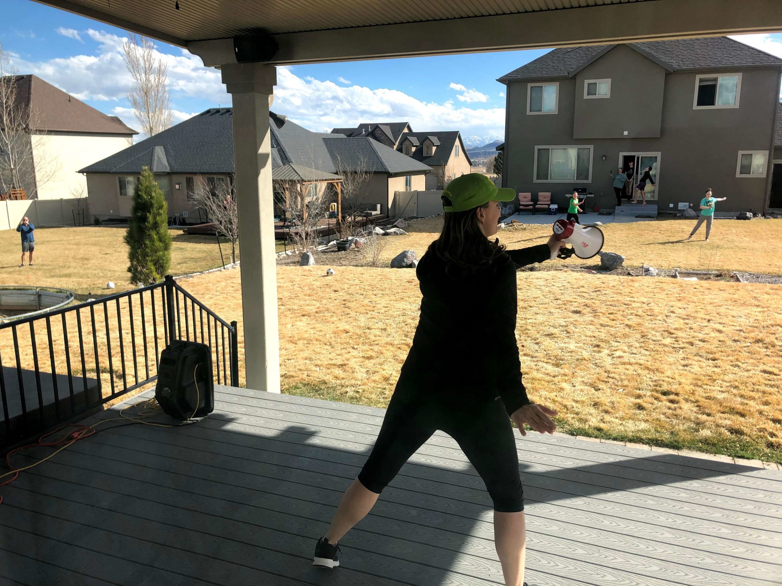 Person holds megaphone on porch, directing a group exercising in a spacious backyard, with several houses in the background.