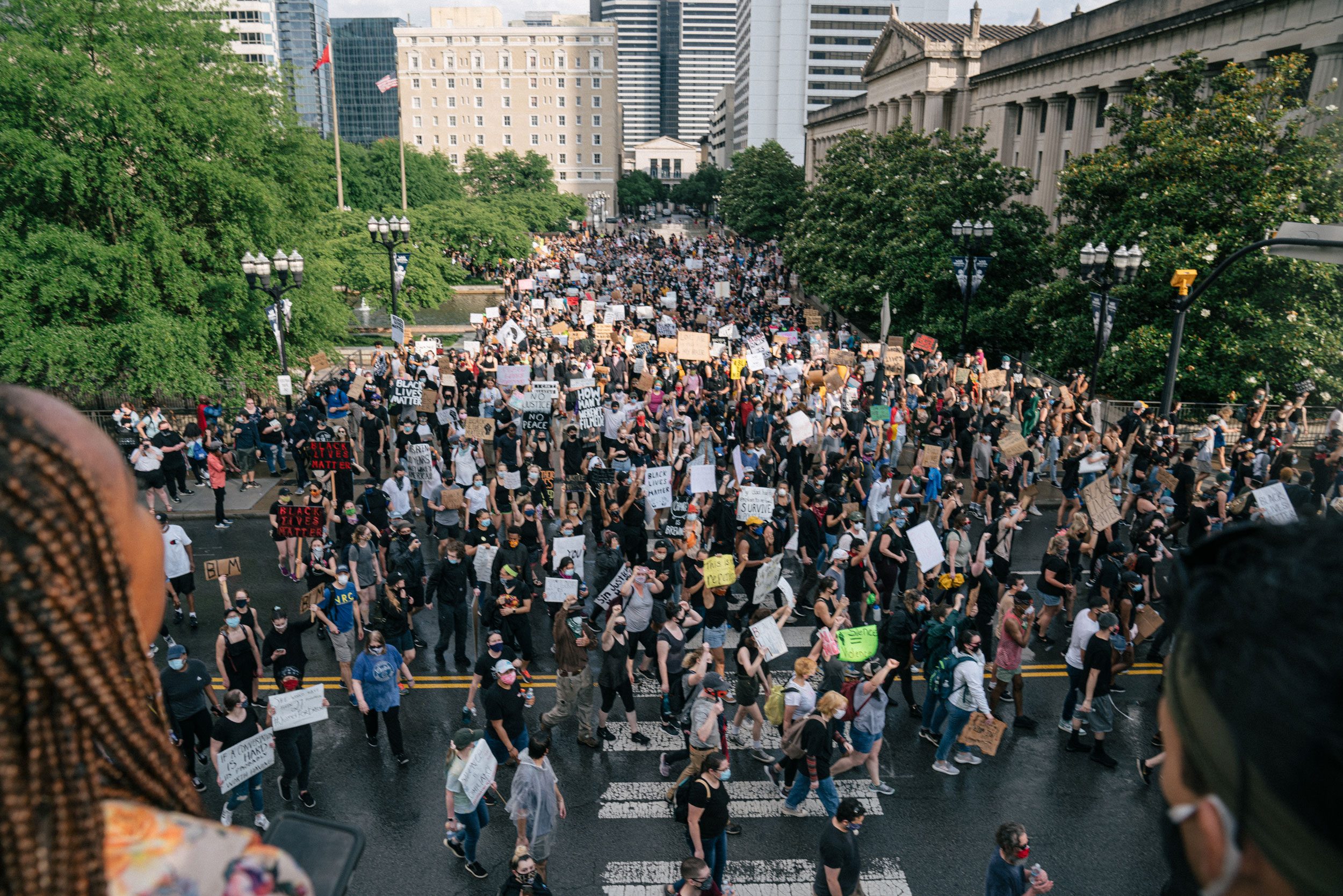 Protesters march holding signs on a city street, surrounded by greenery and buildings.