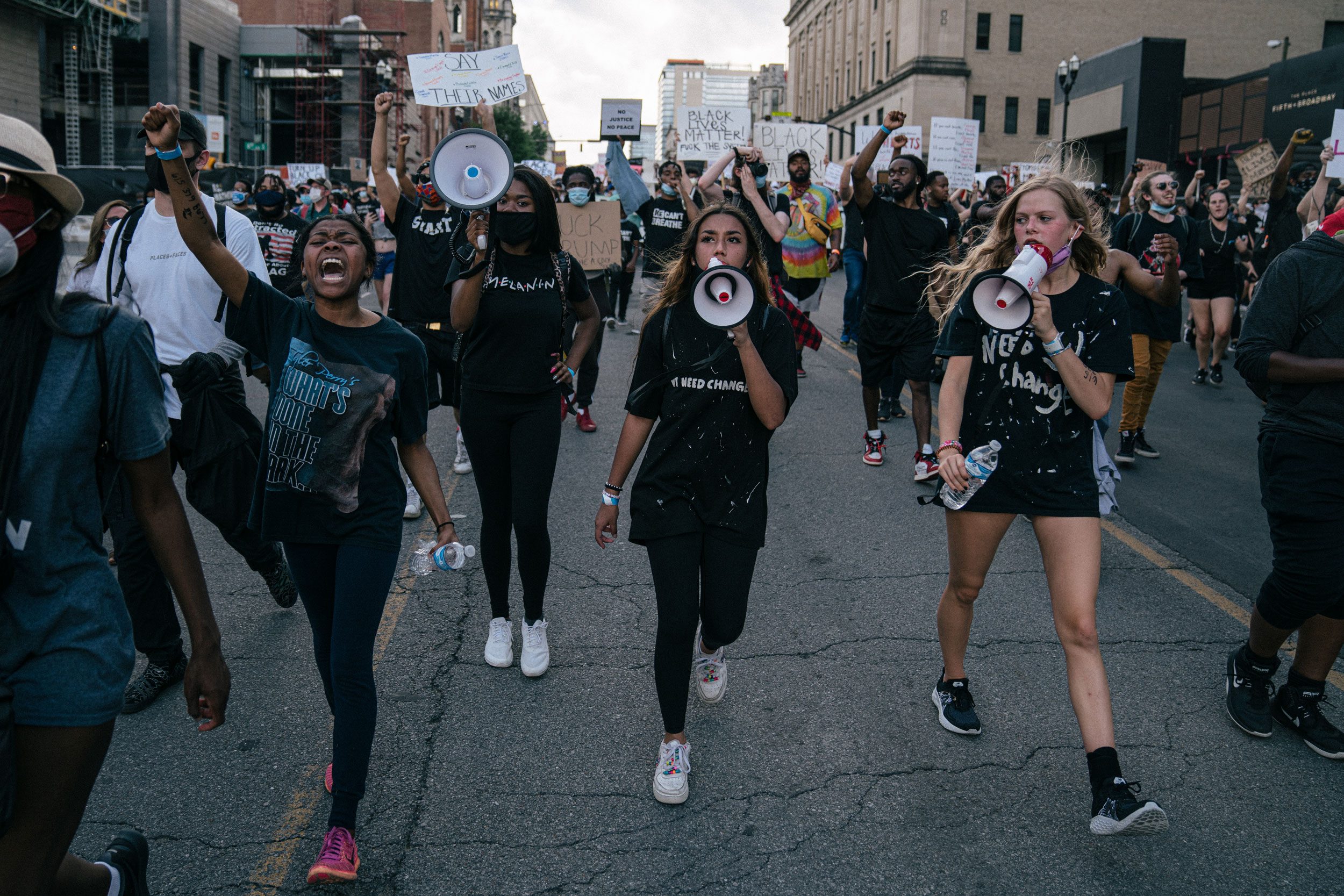 Protesters march down a city street, holding signs and shouting through megaphones, surrounded by buildings and other demonstrators.