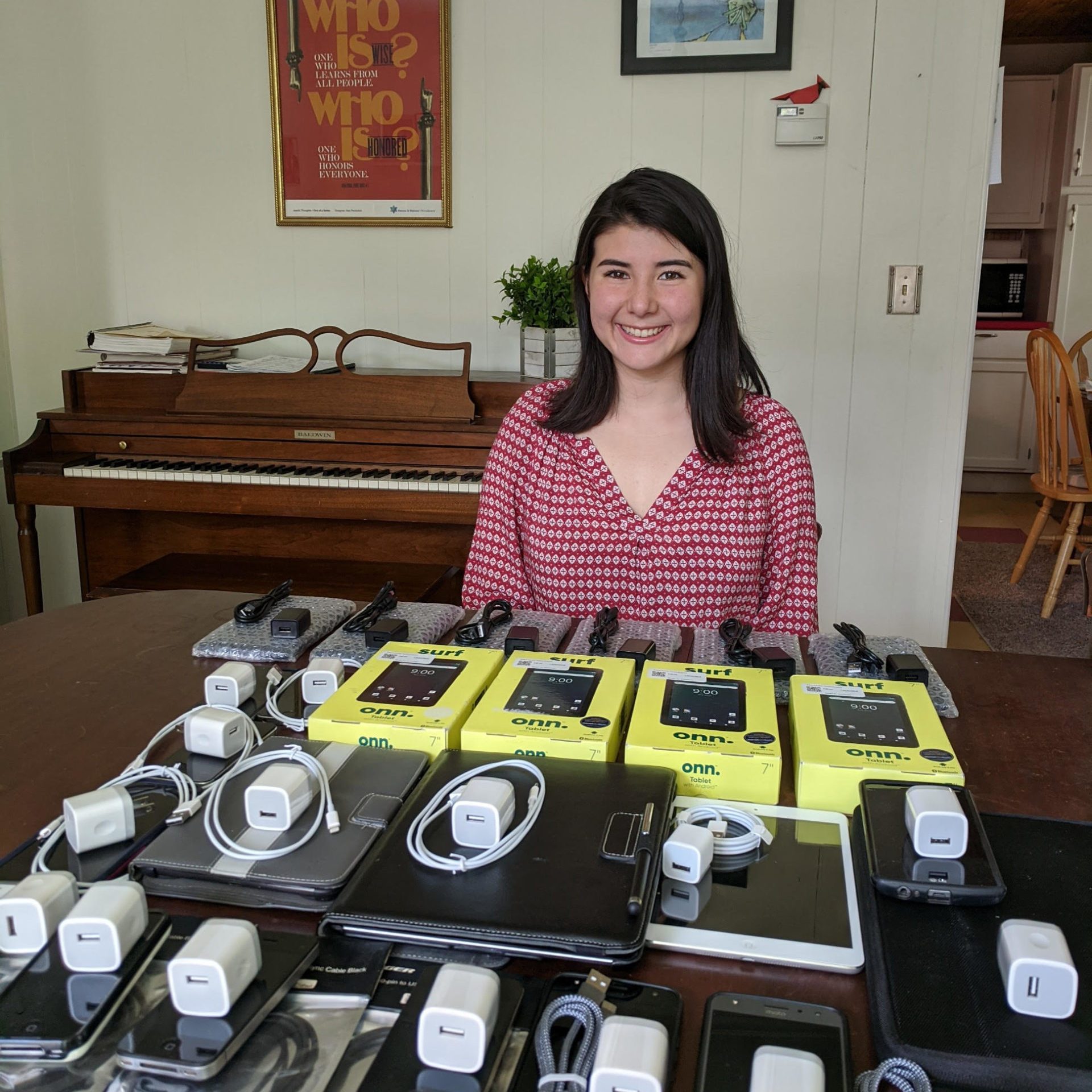 A person sits at a table displaying electronic devices and chargers, framed by a piano and posters in a cozy room.