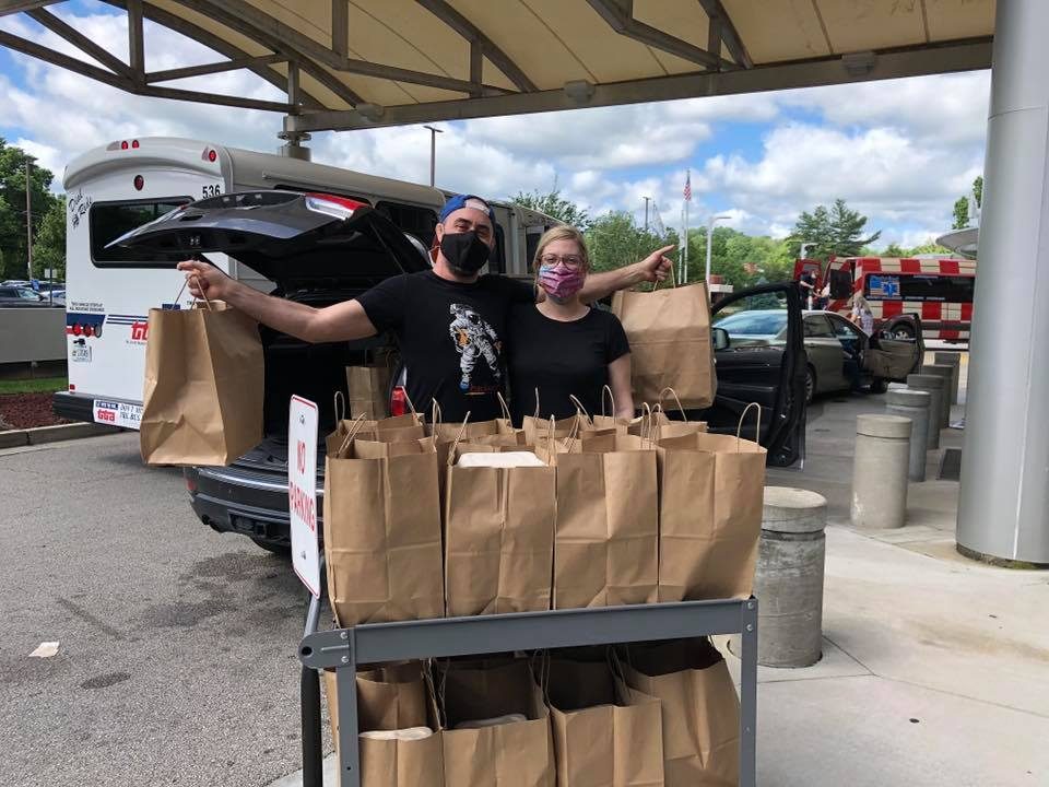 Two people, holding paper bags, stand near a cart full of bags outside, next to a vehicle with a bus and van nearby.