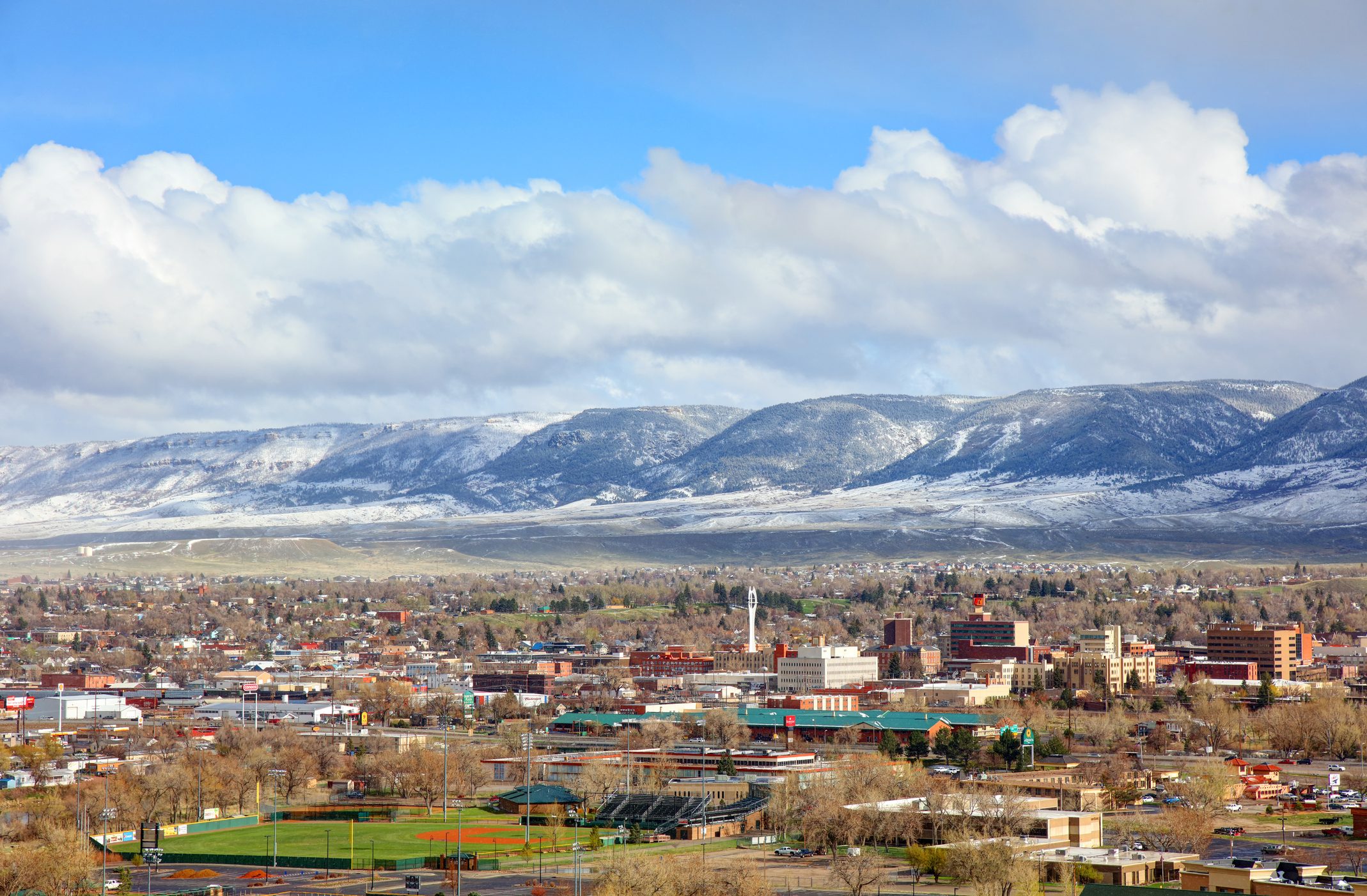 Snow-capped mountains stand still behind a sprawling cityscape, surrounded by baseball fields and distant clouds under a bright blue sky.