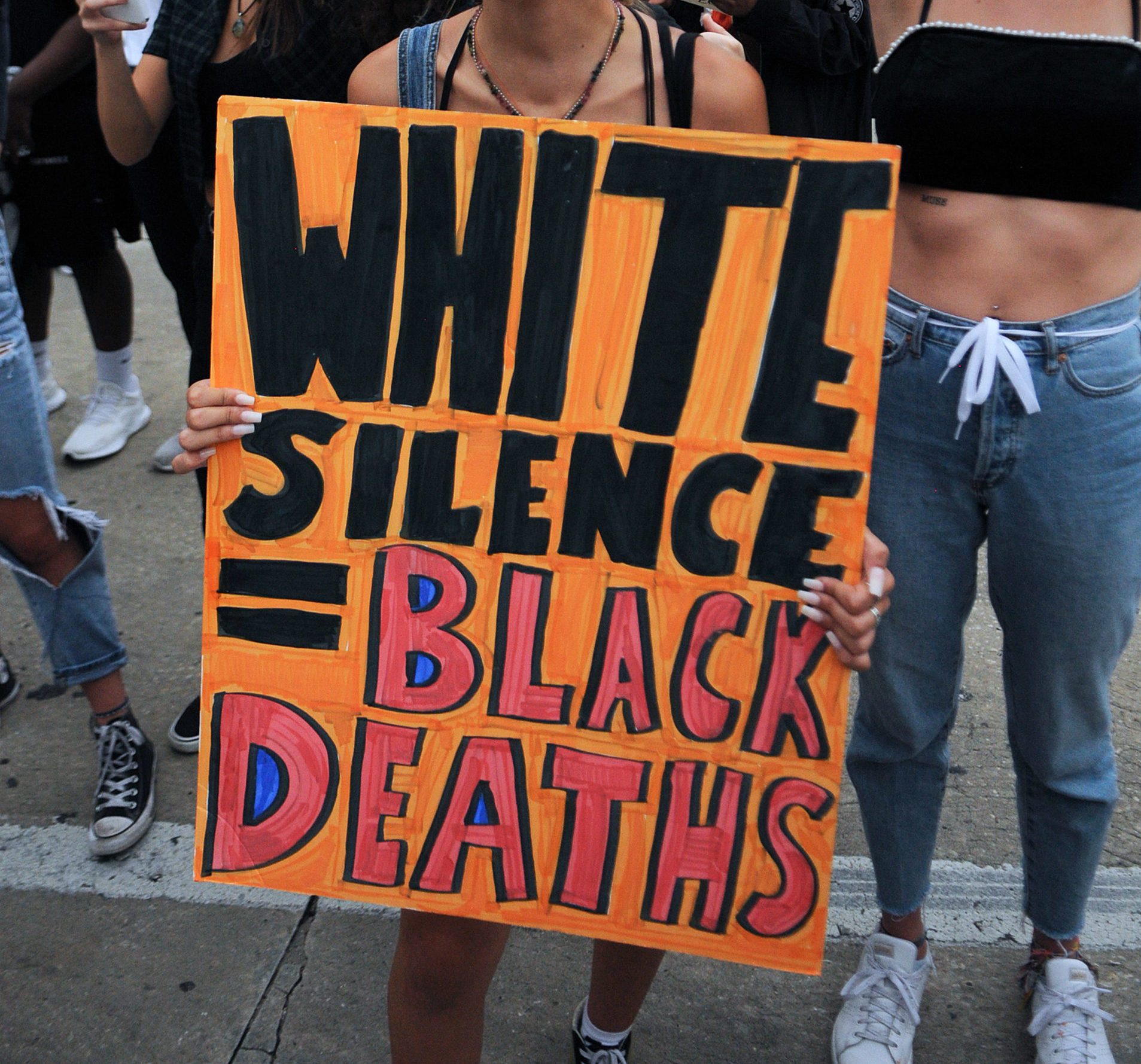 Sign displays "WHITE SILENCE = BLACK DEATHS," held by a person among a group, standing on a city street.