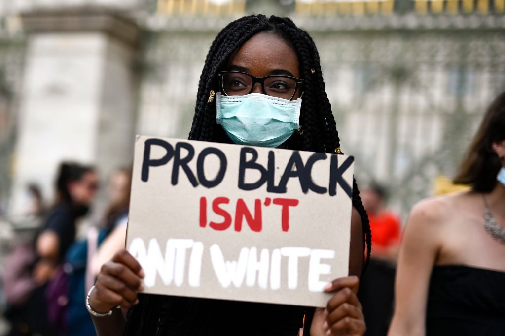 A person holds a sign reading "PRO BLACK ISN'T ANTI WHITE" in a public gathering, wearing a mask, surrounded by blurred individuals.