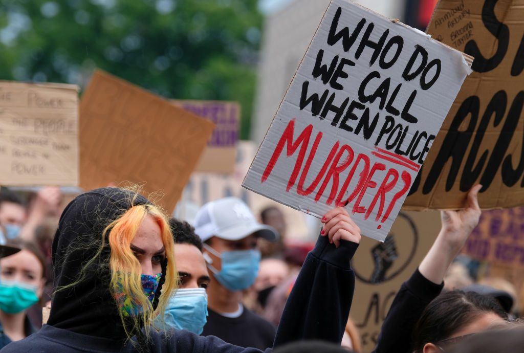 Protesters hold signs, including â€œWho do we call when police murder?â€, in a demonstration with trees and other people in the background.
