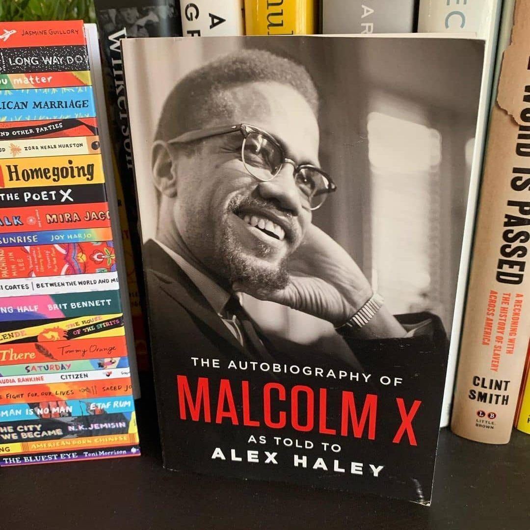 Book features Malcolm X smiling on the cover, titled "The Autobiography of Malcolm X" by Alex Haley, surrounded by colorful books on a shelf.