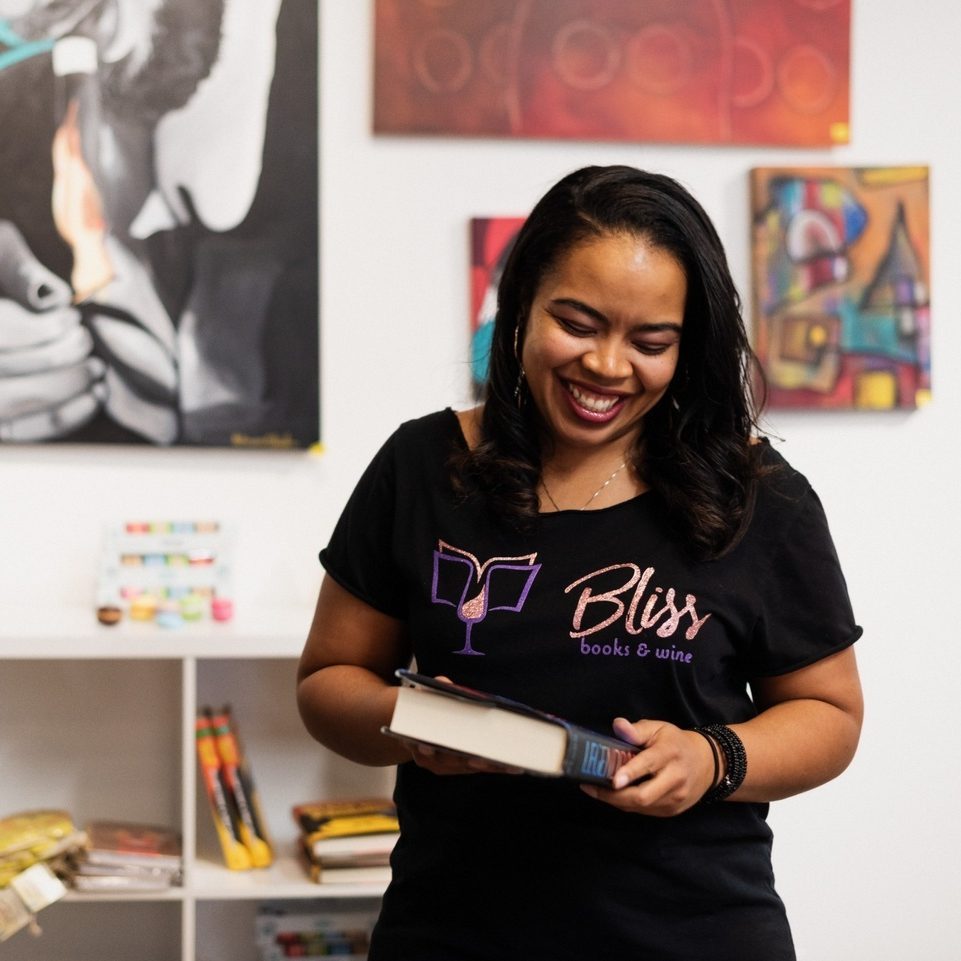Woman holds book, smiling in art-decorated room. Wearing "Bliss books & wine" shirt with colorful, abstract paintings on the wall behind her.